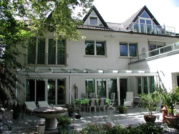 Großes Einfamilienhaus mit moderner Glasüberdachung, Terrasse und Gartenmöbeln im Grünen.