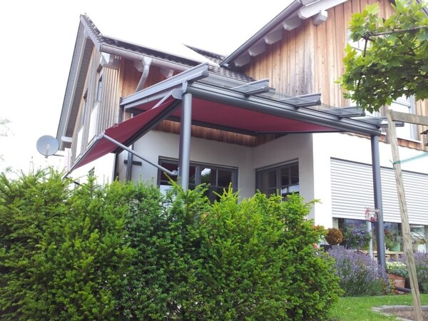Moderne Terrassenüberdachung mit rotem Sonnenschutz an einem Einfamilienhaus im Garten.